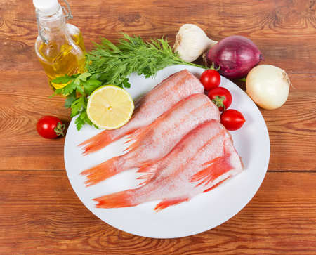 Raw headless gutted carcasses of redfish also known as ocean perch on the dish and vegetables on the rustic table, top view
の写真素材