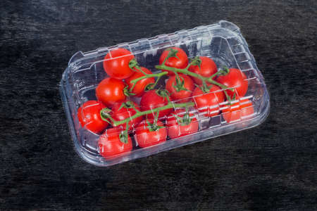 Red cherry tomatoes on branches in the closed plastic sale packaging in the form of the disposable transparent food container on the black surface
の写真素材
