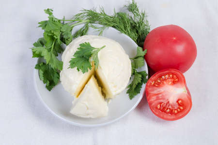 Partly cut  of ball of soft cheese taken out from a brine on the saucer, fresh tomatoes, dill and...