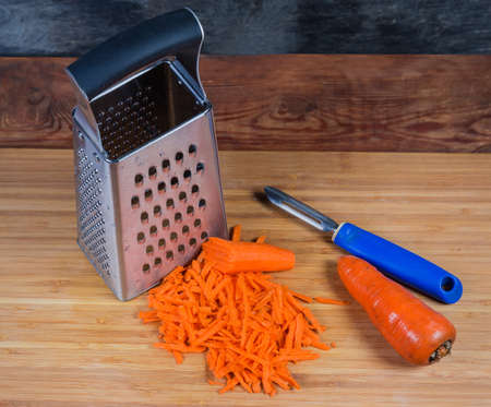 Stainless steel box-shaped kitchen grater, vegetable peeler, grated carrot in large slices and uncleaned whole carrot on the wooden cutting boardの写真素材