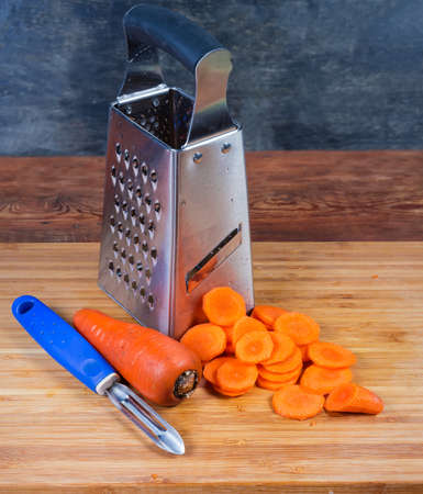 Stainless steel kitchen grater, vegetable peeler, whole uncleaned carrot and carrots sliced into...