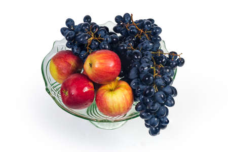 Washed clusters of ripe blue grapes and red apples covered with water drops in the vintage glass vase for fruits on a leg on a white backgroundの写真素材