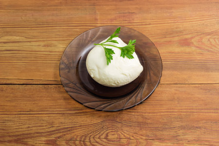 Small ball of brined Mozzarella cheese decorated with parsley twig on a dark glass saucer on a rustic tableの写真素材