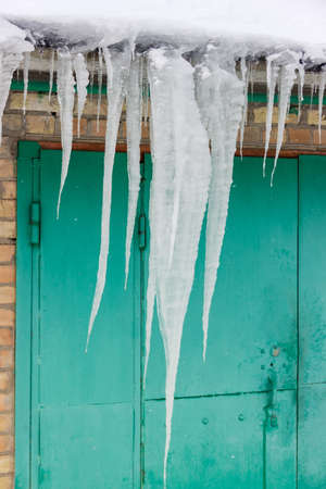 Several long icicles hanging from the snowy roof of industrial building against the gate and part of the brick wall in cloudy wheaterの写真素材