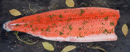 Chilled salmon fillet on the skin sprinkled with chopped dill lies on a dark surface among the...
