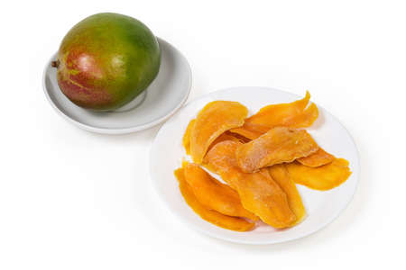 Dried slices of mango pulp on white dish against a whole fresh mango on saucer on a white backgroundの写真素材