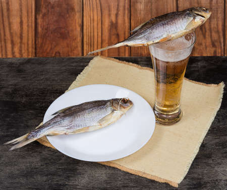 Two salted and air-dried roach fish lie on a beer glass with lager beer and on dish on a napkin on a dark tableの写真素材