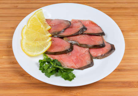 Slices of smoked pink salmon on skin with lemon slices and parsley twig on dish on a wooden surfaceの写真素材