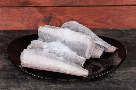 Uncooked frozen carcasses of Argentine hake, or hake hubbsi without the heads and tails covered with hoarfrost on a black dish on dark tableの写真素材
