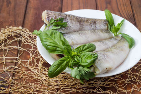 Several raw carcasses of the notothenia fish without of heads and tails and twigs of greens on dish on a rustic table with fishing net, close-upの写真素材