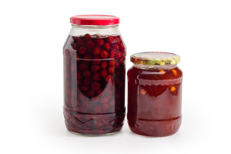Apricot jam with whole apricot kernels addition and cherry jam with whole berries without stones in two glass jars on a white backgroundの写真素材
