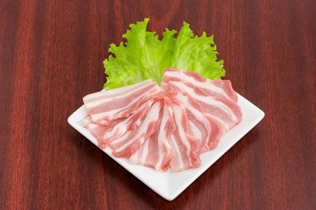 Slices of uncooked streaky bacon with lettuce on a white square dish on a wooden tableの写真素材