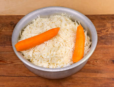 Shredded white cabbage and peeled whole carrots for preparing sauerkraut in large metal mixing bowl on a rustic table, top viewの写真素材