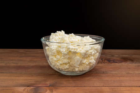Cottage cheese in the transparent glass bowl on the rustic table, side view on a dark backgroundの写真素材
