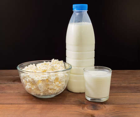 Cottage cheese in the transparent glass bowl, plastic bottle and glass of milk on the rustic table, side view on a dark backgroundの写真素材