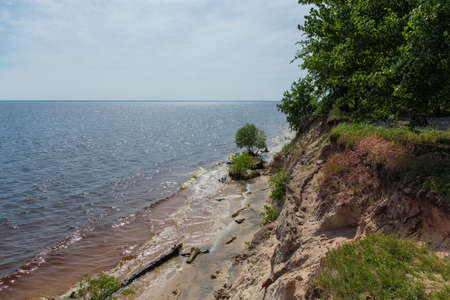 Steep precipitous sandy shore of big reservoir overgrown with forest against the sky with light...