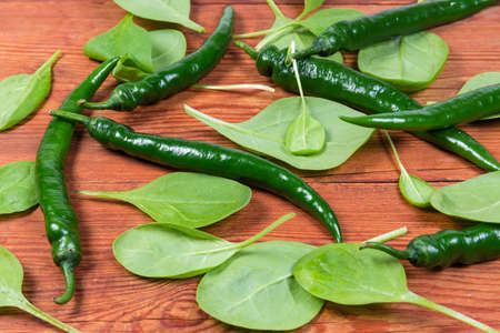Fresh spinach leaves and green chili pods scattered on the old rustic table, fragment close-up