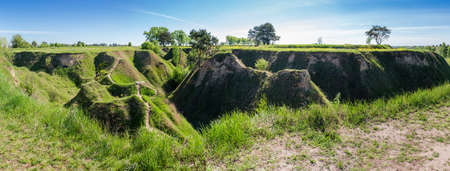 Deep narrow branched ravine with steep precipitous clay slopes overgrown with grass and single trees among flat terrain in springtime, panoramic viewの写真素材