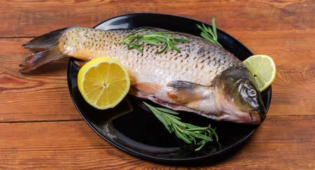 Raw gutted carcass of the carp cleaned from scales and prepared for cooking, fresh rosemary twigs and lemon on black dish on the old rustic tableの写真素材