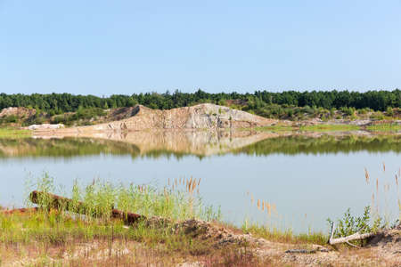 Shallow lake on the site of the abandoned quarry of ilmenite ore with dumps on a opposite bank against the forest and clear sky in summer dayの写真素材
