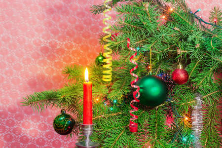 Branches of the Christmas tree, decorated with Christmas ornaments and lights and burning red candle in a candlestick on a blurred red backgroundの写真素材