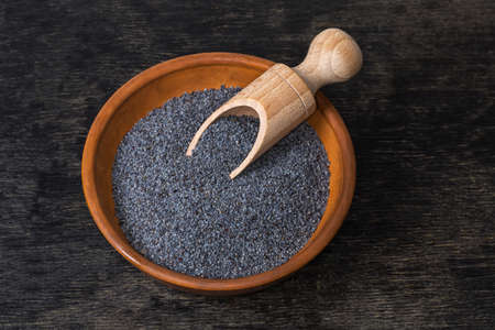 Dry raw poppy seeds of blue variety in the clay bowl with wooden spice spoon on a black surfaceの写真素材