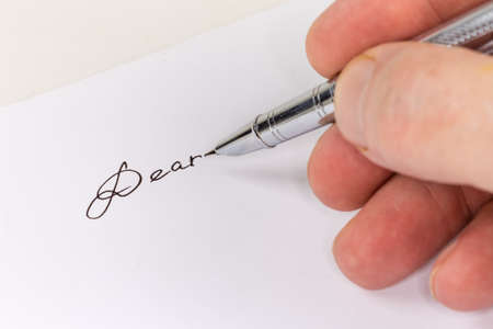 Modern metal fountain pen in hand above the white paper sheet with word salutation "Dear" written by black ink, close-up in selective focusの写真素材