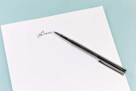 Modern metal fountain pen lie on a white paper sheet with word salutation "Dear" written by black ink on a blue surfaceの写真素材