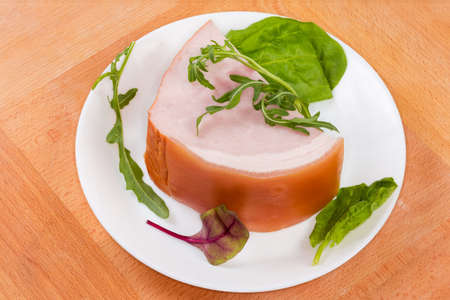 Piece of boiled smoked ham on skin among the leaves of greens on the white dish on the wooden surfaceの写真素材