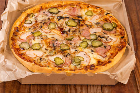 Round pizza with different ham, mushrooms and pickled cucumbers served in open cardboard pizza box on the old rustic tableの写真素材