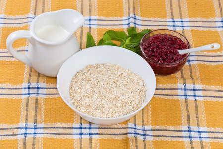 Raw thin-rolled oats in the white bowl, hot milk in milk jug and red currants jam on a table with tableclothの写真素材