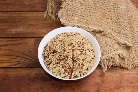 Mixture of the raw long-grained brown, red and black rice in a white bowl on the wooden rustic table...