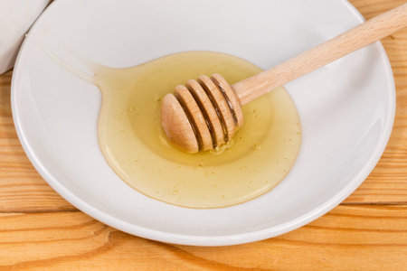 Fresh liquid light honey on the saucer with a wooden honey dipper on the rustic table, close-upの写真素材