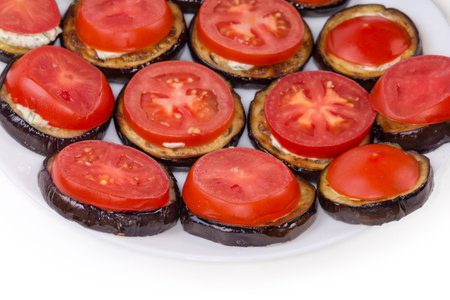 Roasted chopped by circles eggplants with garlic sauce and covered by cut circles of fresh tomatoes on dish, fragment on a white backgroundの写真素材