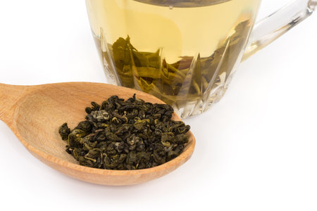 Dried green tea leaves in the wooden spoon against the brewed green tea in the transparent glass cup, fragment close-up in selective focus on a white backgroundの写真素材