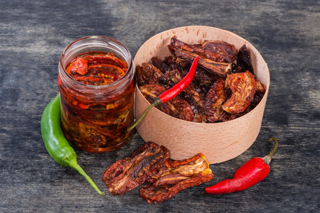 Dry-cured red tomatoes in the round wooden box and next to her, sun-dried tomatoes preserved in cooking oil in jar and red and green chili on the black surfaceの写真素材