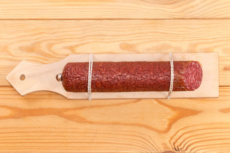 Piece of the dry-cured sausage tied to the specially designed narrow wooden cutting board for...