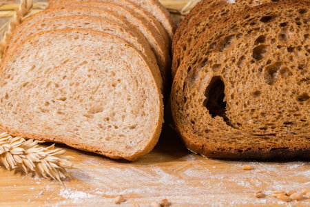 Sliced two oblong loafes of the dark brown bread made from rye and wheat flour and bread with bran among the ears and grain on the cutting board strewn with flour, close-up in selective focusの写真素材