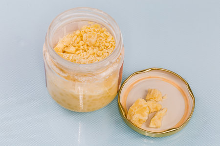 Raw partially crumbled solid cocoa butter in the small glass jar and pieces on a removed and inverted lid on a blue surfaceの写真素材