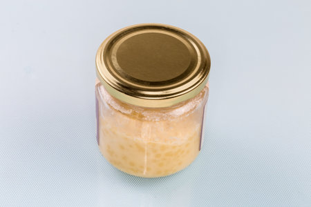 Raw solid cocoa butter in the small glass jar with closed lid on a blue surfaceの写真素材