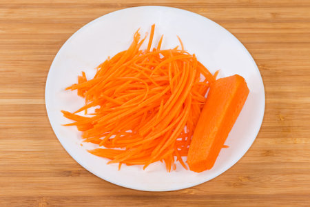 Fresh carrots chopped into thin long slices for spicy Korean carrot salad preparation on a white dish on the wooden surfaceの写真素材