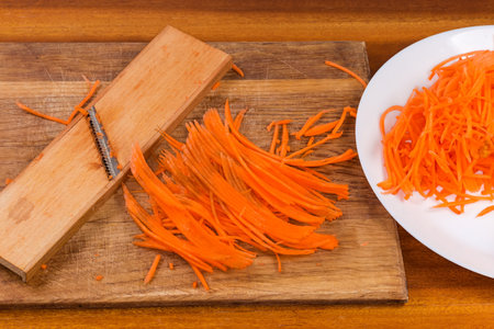 Fresh carrots chopped into thin long slices for spicy Korean carrot salad preparation on a cutting board and on dish and special grater on the wooden surfaceの写真素材