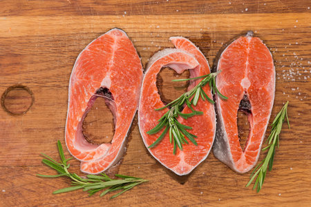Big pieces of the fresh uncooked rainbow trout sprinkled with coarse salt and twigs of the fresh rosemary on the wooden cutting board, top viewの写真素材