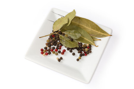 Dried whole bay leaves, allspice and mixture of various whole peppercorns on the square saucer on a white backgroundの写真素材