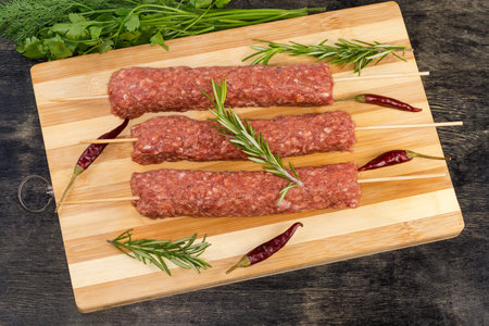 Several raw lula kebab skewered on wooden skewers on the wooden cutting board among the some vegetables, spices and greens on a black surfaceの写真素材