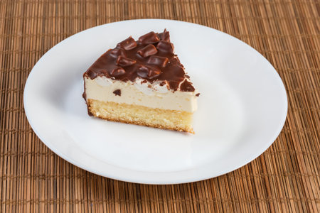 Slice of round layered sponge cake with souffle, marshmallow pieces and chocolate coating on a white dish on a bamboo place matの写真素材