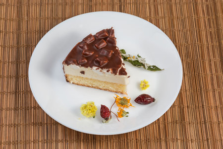 Slice of layered sponge cake with souffle, marshmallow pieces and chocolate coating on a white dish among dried flowers and berries for herbal tea preparation on a bamboo place matの写真素材