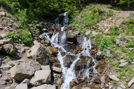 Waterfall on a mountain stream flowing down the slope overgrown with forest in spring sunny dayの写真素材