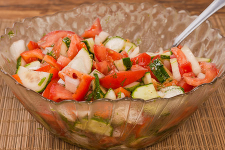 Vegetable salad made with fresh sliced tomatoes, red bell peppers, cucumbers and onion in the glass salad bowl with spoon on the bamboo place mat, close-up in selective focusの写真素材