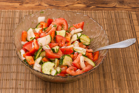 Vegetable salad made with fresh sliced tomatoes, cucumbers and onion in the glass salad bowl with spoon on the bamboo place matの写真素材